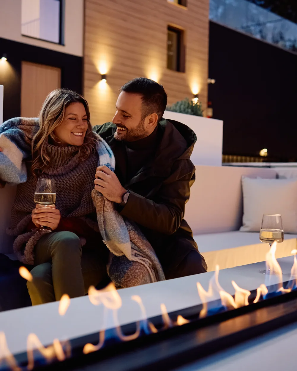 A couple relaxing under a blanket by a lit linear fire table on a modern backyard patio at dusk